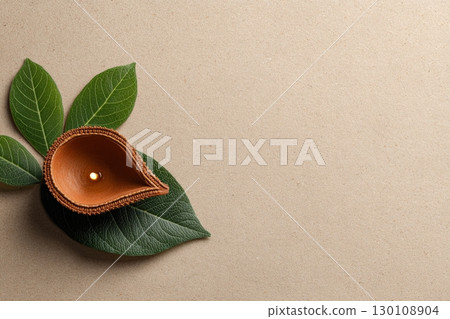 A small clay diya with a lit flame sits on green leaves on a textured beige surface. 130108904