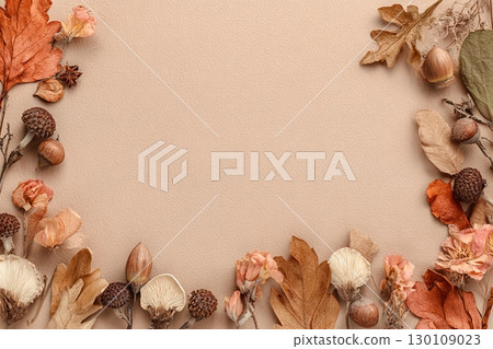 A flat lay of autumn leaves, acorns, and dried flowers arranged around the border of a light brown textured background. 130109023