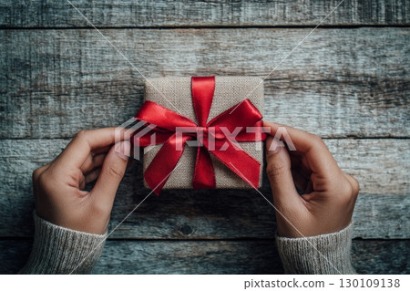 A person holding a small gift wrapped in brown fabric with a red ribbon on a wooden table. 130109138