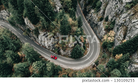 A winding road cuts through a dense forest with a red car driving along it, surrounded by trees and rocky slopes. 130109255