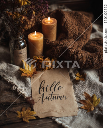 A cozy autumn setting with lit candles, a 'Hello Autumn' card, dried flowers, and colorful leaves on a knitted blanket. 130109312