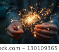 Woman holding a lit sparkler with both hands, creating a warm glow. 130109365
