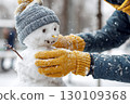 A person is putting the finishing touches on a small snowman wearing a hat in snowy weather. 130109368
