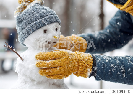 A person is putting the finishing touches on a small snowman wearing a hat in snowy weather. 130109368