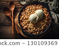A warm apple crumble topped with two scoops of vanilla ice cream in a round dish, placed on a wooden table with two wooden spoons and a cloth nearby. 130109369