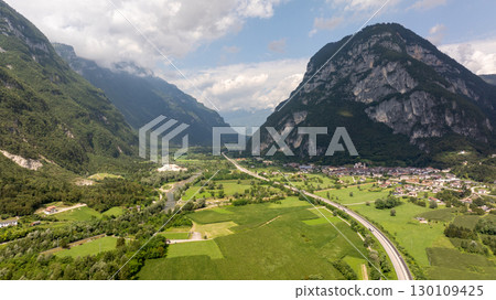 Aerial view of Valsugana in Italy, showcasing the highway, green fields, river, and mountains under a cloudy sky 130109425