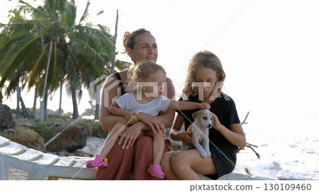 Family of three play with puppy on beach at sunset sharing warm joyful moments together  130109460
