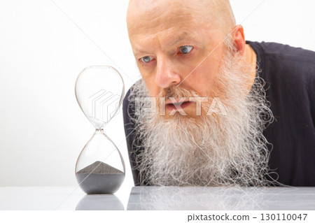 bald man with a white beard closely examines an hourglass bald man with a white beard closely examines an hourglass 130110047