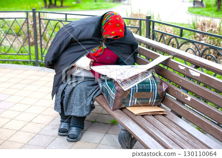 A homeless person in a red headscarf and black coat sits on a park bench, reading a newspaper. A homeless person in a red headscarf and black coat sits on a park bench, reading a newspaper. 130110064