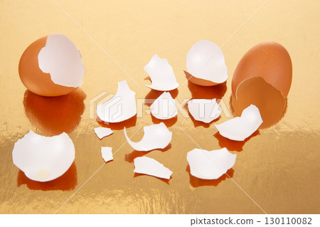 Two broken brown eggs with scattered eggshell fragments on a reflective golden surface 130110082