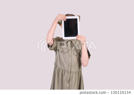 Young caucasian female child holding tablet to face in green dress with neutral Young caucasian female child holding tablet to face in green dress with neutral 130110134