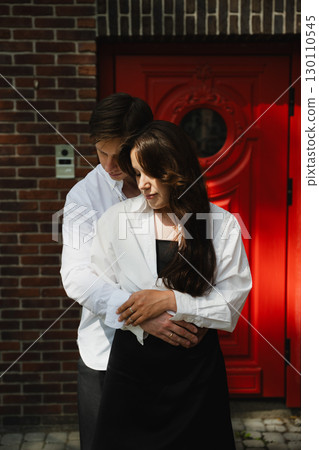 Young loving married couple embracing in front of a vibrant red door with brick wall background 130110545