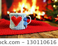 a mug in a knitted case with a red heart on the side on a wooden table against the background of a red plaid, a fireplace and a Christmas tree 130110616