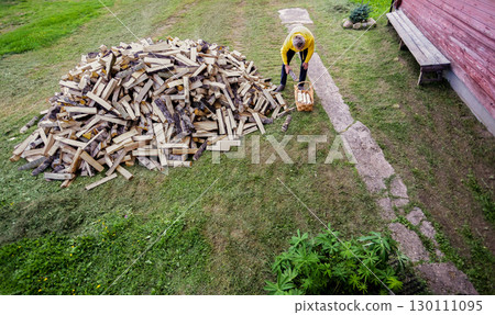 Gray haired man takes firewood from pile near country house and puts it into large wicker basket. 130111095