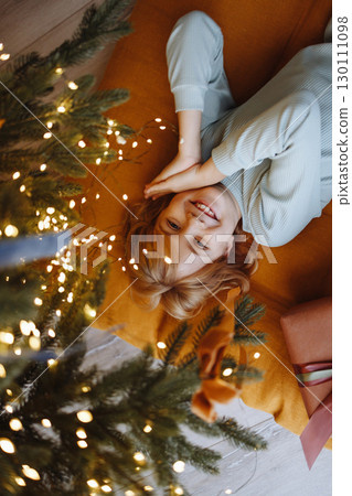 Smiling child top view under Christmas tree with gift box on orange cushion on wooden floor 130111098
