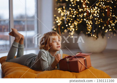 Smiling girl in pajamas holding gift box lying on cushion near Christmas tree with lights 130111100