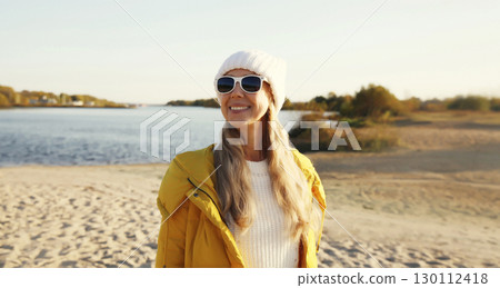 Happy woman standing on sunny beach looking away, wear jacket, sea coast 130112418