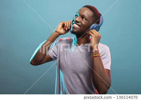 Cheerful male individual looking up and enjoying his favorite songs against a blue background. Happy african american man in casual attire, touching his wireless headphones and listening to music. Cheerful male individual looking up and enjoying his favorite songs against a blue background. Happy african american man in casual attire, touching his wireless headphones and listening to music. 130114070