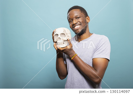 Cheerful black male person stands and grasps a skull close to him, posing for the camera in a studio. Portrait of a happy african american man holding a human skeletal head. 130114097