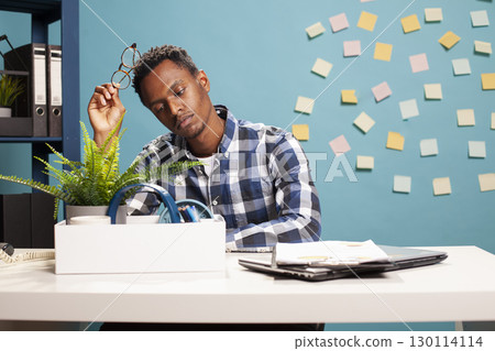 African American manager upset after losing his job due to financial struggles at the company. Unemployed ooffice worker with glasses in his hand, seated at desk, sadly looking at his box of items. African American manager upset after losing his job due to financial struggles at the company. Unemployed ooffice worker with glasses in his hand, seated at desk, sadly looking at his box of items. 130114114
