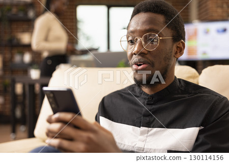 Closeup of black male accountant looking at smartphone with surprised expression, reacting to good news about startup company finances. Shocked businessman with glasses holding his mobile device. Closeup of black male accountant looking at smartphone with surprised expression, reacting to good news about startup company finances. Shocked businessman with glasses holding his mobile device. 130114156