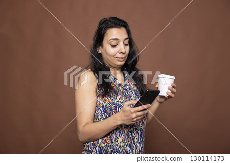 Female individual with disposable cup, using her smartphone, chatting and browsing on social media. Indian woman holding disposable cup and mobile device as she stands against isolated background. 130114173