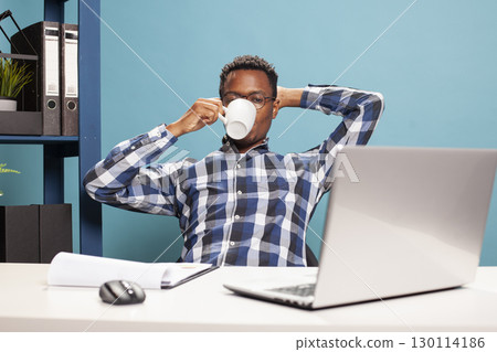 African american male individual relaxing on chair and drinking coffee as he watches a video on his laptop. Young man seated at desk, leaning backwards with a cup in hand and looking at device screen. African american male individual relaxing on chair and drinking coffee as he watches a video on his laptop. Young man seated at desk, leaning backwards with a cup in hand and looking at device screen. 130114186