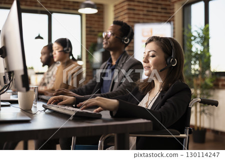 Dedicated female professional in wheelchair, wearing wired headphones, stays focused on tasks at her desk. White woman entrepreneur interacts with online customers, updating information on computer. 130114247