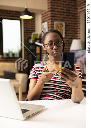 African american businesswoman seated at desk, holding mobile device during virtual meeting with investors. Black female manager working remotely from home office, engaged in professional video call. 130114249