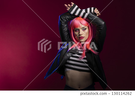 Fashion forward woman with vibrant pink hair and striped crop top poses in dramatic scene. Stylish female model with arms above head, confidently stands in artistic studio lighting. 130114262