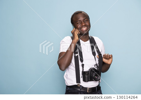 Cheerful black man in suspenders talks on his mobile phone, standing in a blue background studio. He looks at the camera with a natural and friendly expression, embodying modern communication. Cheerful black man in suspenders talks on his mobile phone, standing in a blue background studio. He looks at the camera with a natural and friendly expression, embodying modern communication. 130114264