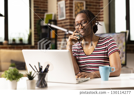 Self employed woman sits at desk, multitasking, speaks with clients on phone and updates data on laptop. Smiling female freelancer uses mobile device while working from home on personal computer. 130114277