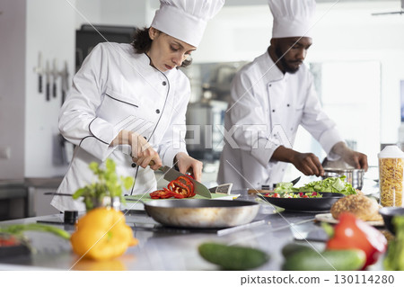 Woman chef in uniform slicing a bell pepper in professional kitchen, using fresh produce and kitchen tools to create a delicious dish. Culinary expert prepared ingredients on the stove. Woman chef in uniform slicing a bell pepper in professional kitchen, using fresh produce and kitchen tools to create a delicious dish. Culinary expert prepared ingredients on the stove. 130114280