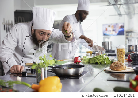 In a restaurant kitchen, chef garnishes a dish with fresh thyme and parsley on the stove. Young culinary expert cooking with colorful vegetables and spices, organic raw materials. In a restaurant kitchen, chef garnishes a dish with fresh thyme and parsley on the stove. Young culinary expert cooking with colorful vegetables and spices, organic raw materials. 130114291