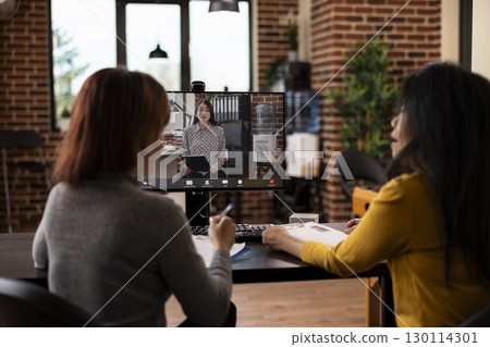 Female colleagues seated at desk, on a video conference call with their asian manager seen on desktop monitor. Businesswomen in office using computer for a virtual meeting with potential partner. 130114301