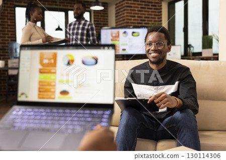 Black businessman seated on couch, holding clipboard and consulting with coworker during office meeting. Two african american professional executives review financial and accounting reports together. Black businessman seated on couch, holding clipboard and consulting with coworker during office meeting. Two african american professional executives review financial and accounting reports together. 130114306