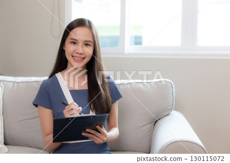 Smiling young Asian female therapist holding a pen and clipboard sits on sofa, expressing warmth, trust and empathy in mental health support session setting. Counseling professional concept. 130115072