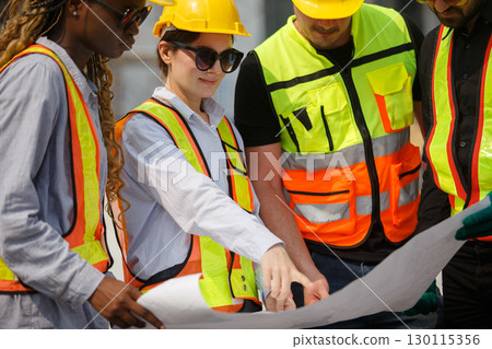 Diverse team of construction workers wearing safety helmets and reflective vests looking blueprint discussing building plans on construction site for teamwork and project planning concepts. Diverse team of construction workers wearing safety helmets and reflective vests looking blueprint discussing building plans on construction site for teamwork and project planning concepts. 130115356