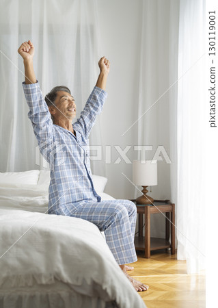 Senior man in pajamas sitting on bed and stretching 130119001