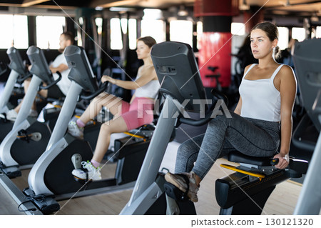 Girl performs exercises with stationary bicycle in gym Girl performs exercises with stationary bicycle in gym 130121320