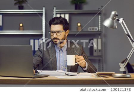Serious, focused young man in glasses, dressed in office style, is sitting at his desk in the office drinking tea or coffee and typing text on laptop. 130121378