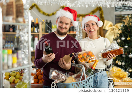 Smiling married couple in santa claus hat with basket in hands choosing food and alcohol in grocery department of supermarket 130121519