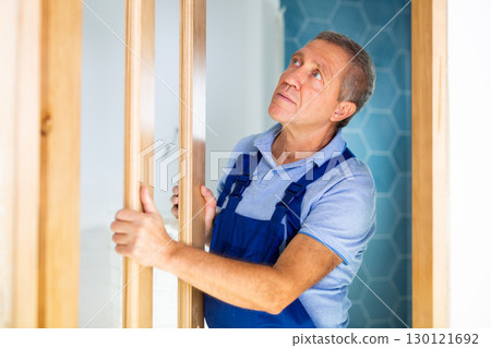 Professional male craftsman in a blue uniform checking the quality of the installed door at the apartment Professional male craftsman in a blue uniform checking the quality of the installed door at the apartment 130121692