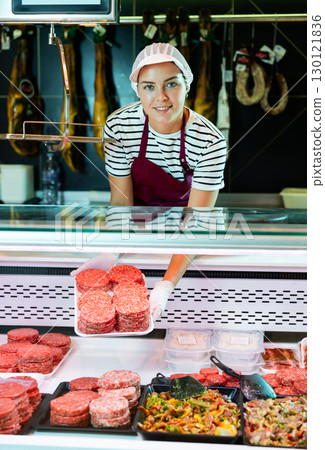 Female butcher shop vendor offering burger patties from display case Female butcher shop vendor offering burger patties from display case 130121836