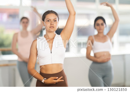 Portrait of pretty young girl dancer exercising various ballet moves during training with group of sporty people in training room 130121850