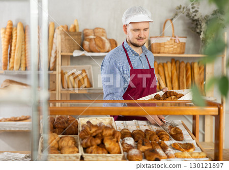 Man employee puts croissants in window, arranges display of goods at bakery. Man employee puts croissants in window, arranges display of goods at bakery. 130121897