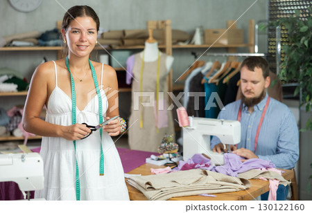 Young female dressmaker welcoming clients while man is sewing behind 130122160