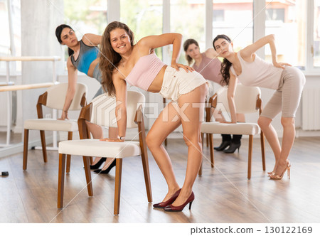 Leaning on chair seat with hand foot, young woman performs lap dance during group workout. 130122169