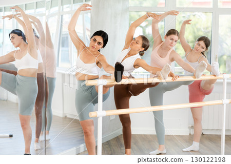 Group of fit young female ballet dancers learn grand batman position near ballet barre in gym Group of fit young female ballet dancers learn grand batman position near ballet barre in gym 130122198