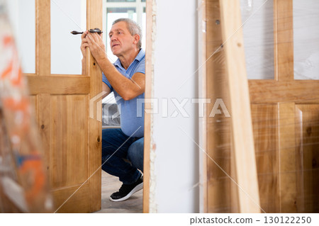 Professional middle-aged repairman installing a door handle on a door during renovation, indoors Professional middle-aged repairman installing a door handle on a door during renovation, indoors 130122250